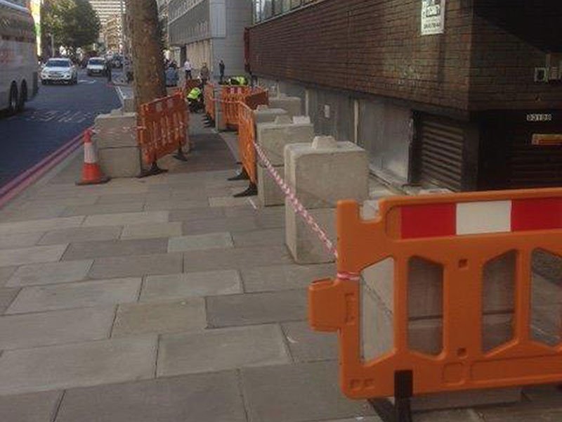 Kentledge blocks lining a city pavement behind orange pedestrian barriers