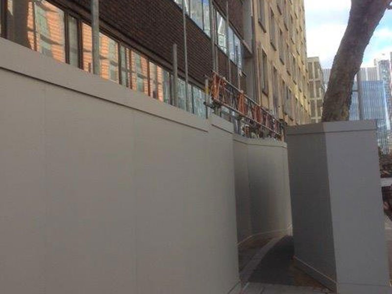 Tall site hoarding ballasted by concrete blocks running along a city street