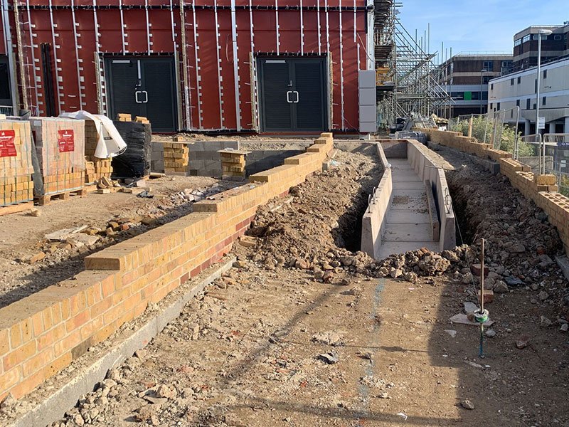 Concrete cable troughs bedded into a trench beside a building under construction