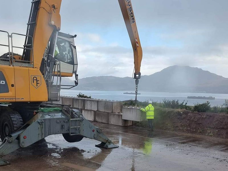 Excavator placing large interlocking blocks on a wall beside a sea loch