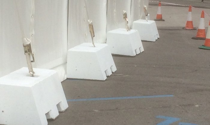 Counterweight blocks anchoring marquee guy lines on a hardstanding beside traffic cones