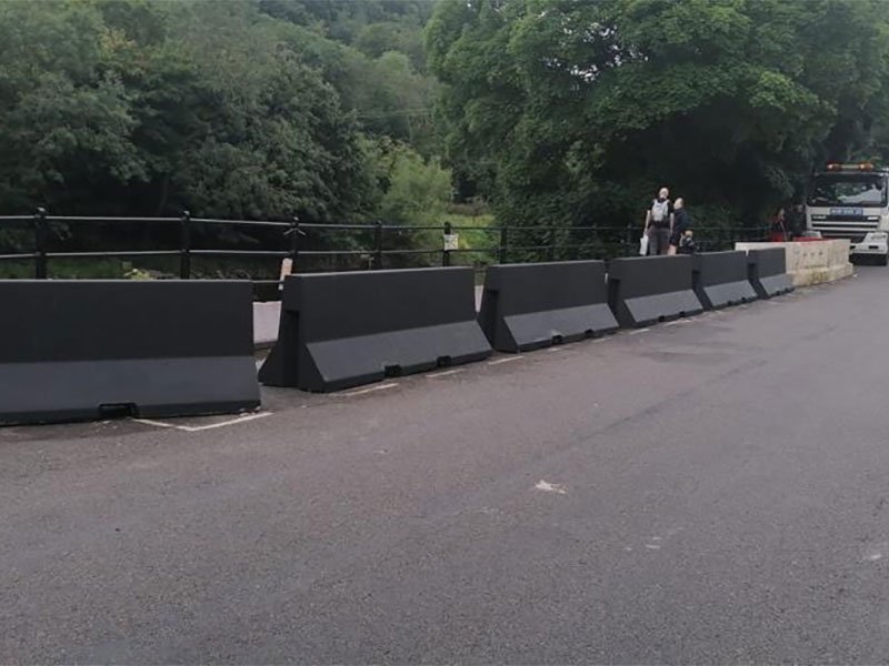 Black-coated Jersey barriers lining a road behind steel railings