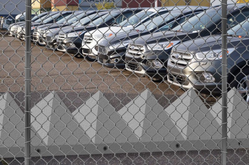 Row of pyramid tank trap barriers behind a chain link fence at a car compound