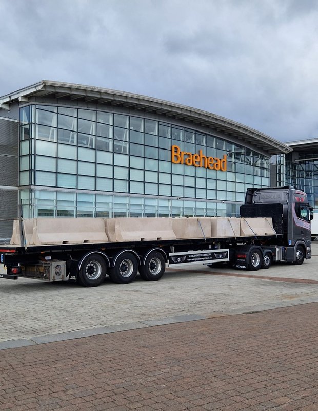 Lorry loaded with Jersey barriers parked outside a glass-fronted retail centre