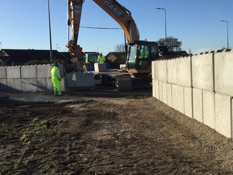 Excavator installing a Legato block wall along a roadside with banksmen in hi-vis