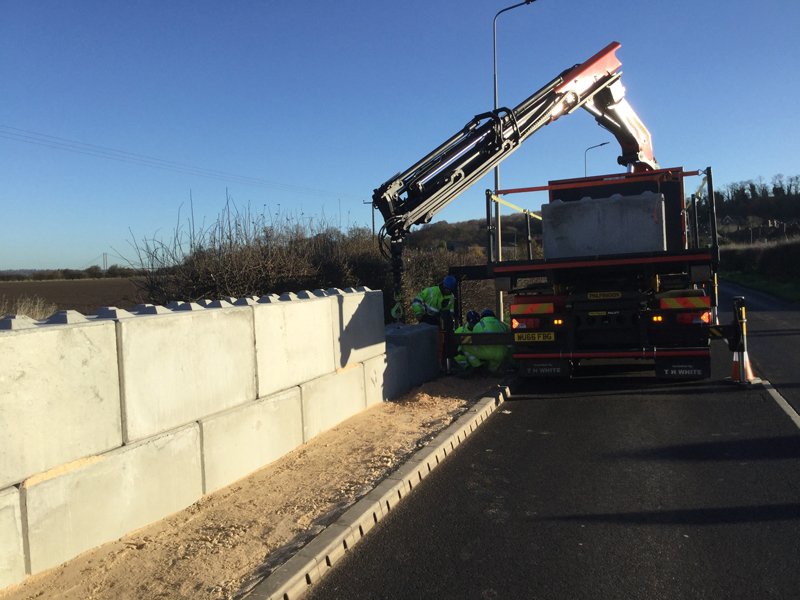 Lorry-mounted crane lifting a Legato block onto a roadside flood wall