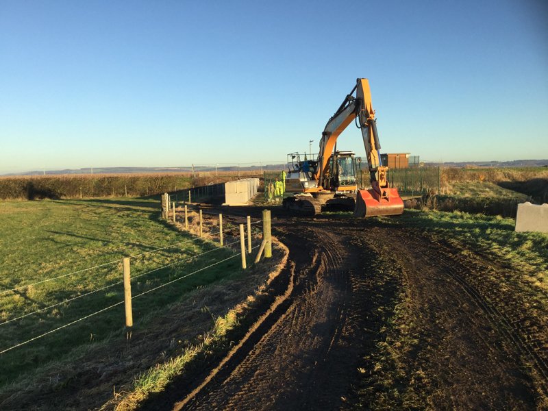 Excavator building an interlocking block wall along a muddy field track