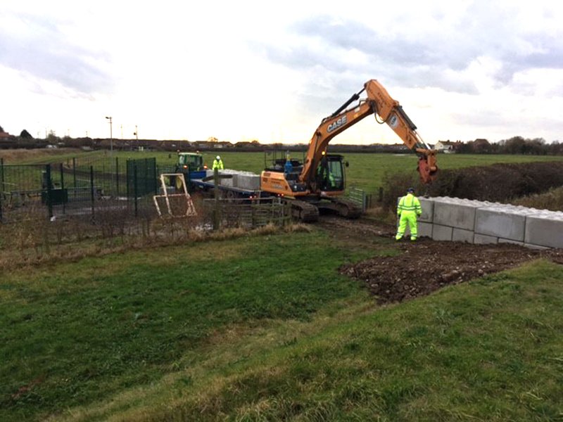 Excavator placing interlocking blocks for a flood defence wall in a field