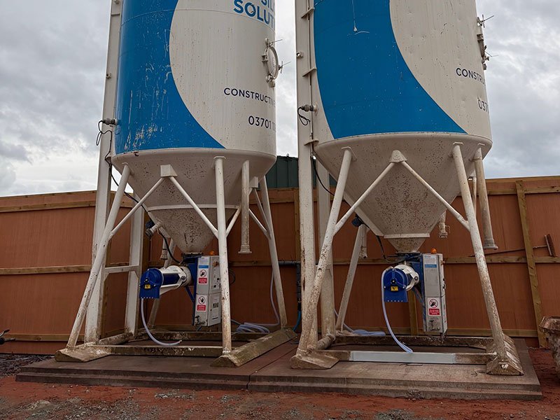 Two site silos standing on precast concrete foundation slabs behind orange hoarding
