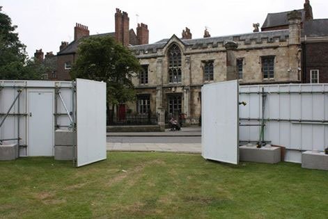 Kentledge blocks ballasting white hoarding panels on grass by a historic building