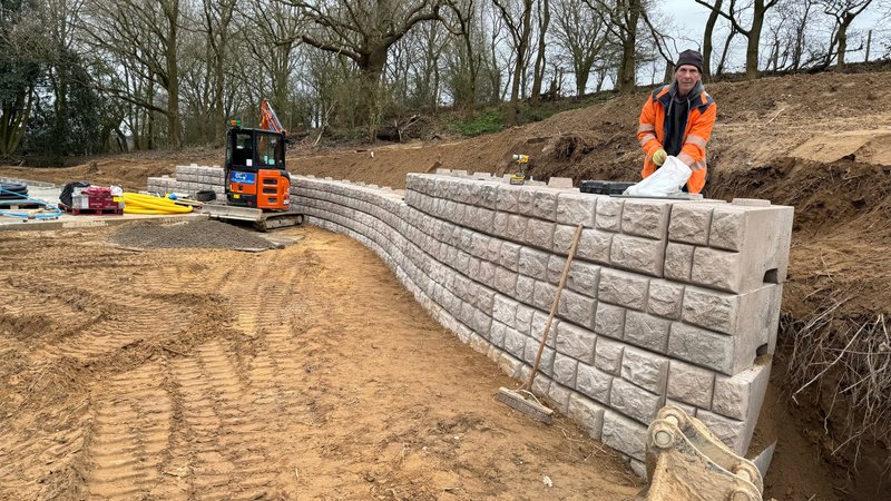 Worker installing curved cobblestone-finish Retainer-Rock units with a mini excavator