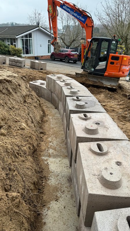 Excavator setting Retainer-Rock walling units into a trench beside homes