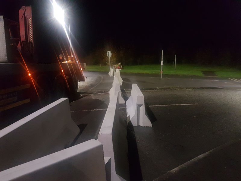 Lorry offloading Jersey barriers across a road at night under floodlights
