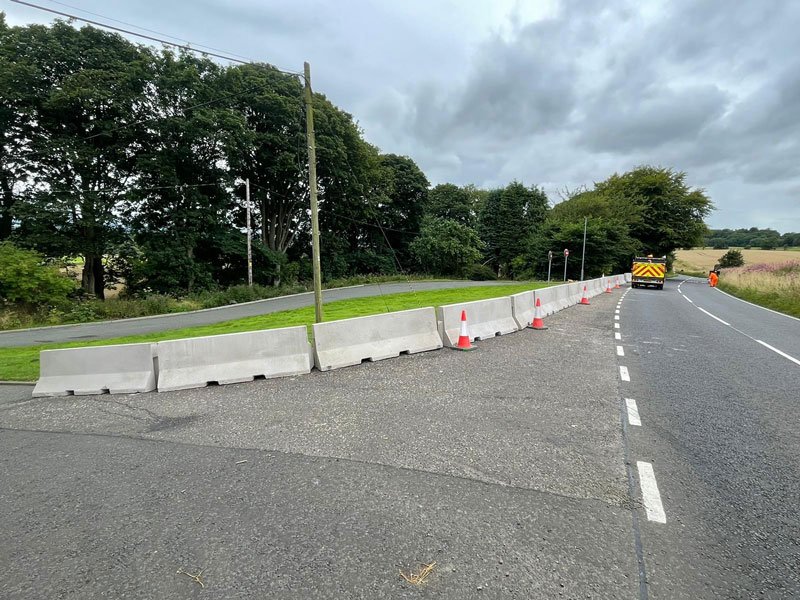 Line of Jersey barriers and cones narrowing a tree-lined rural road