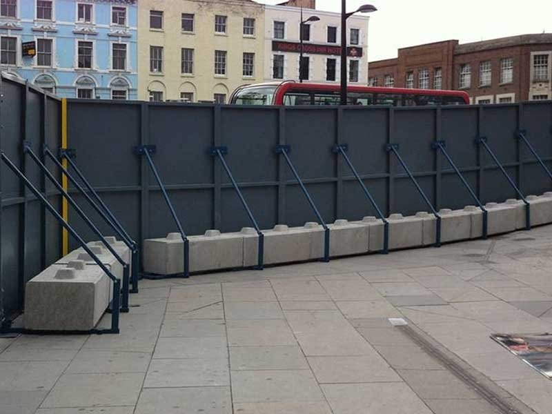 Concrete ballast blocks bracing a dark site hoarding across a city square