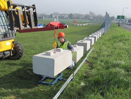 Worker guiding a ballast block into place with a telehandler during fence installation