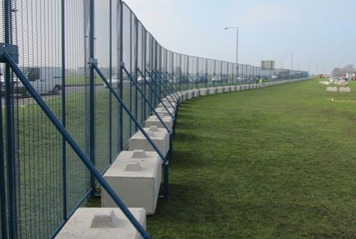 Tall mesh security fencing weighted by ballast blocks curving across grass