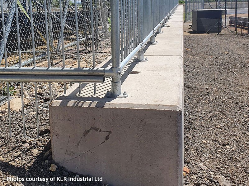 Low concrete retaining wall carrying a perimeter security fence above gravel