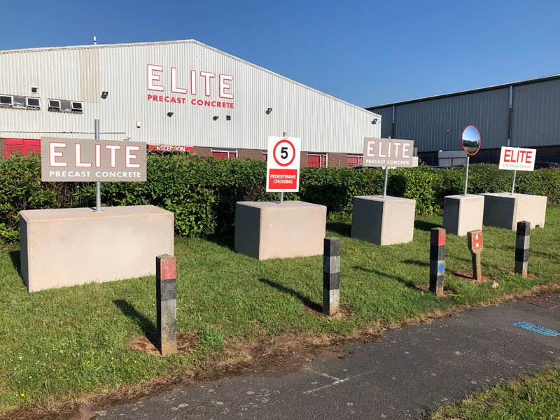 Mobile post blocks holding signage boards along a grass verge at a works entrance