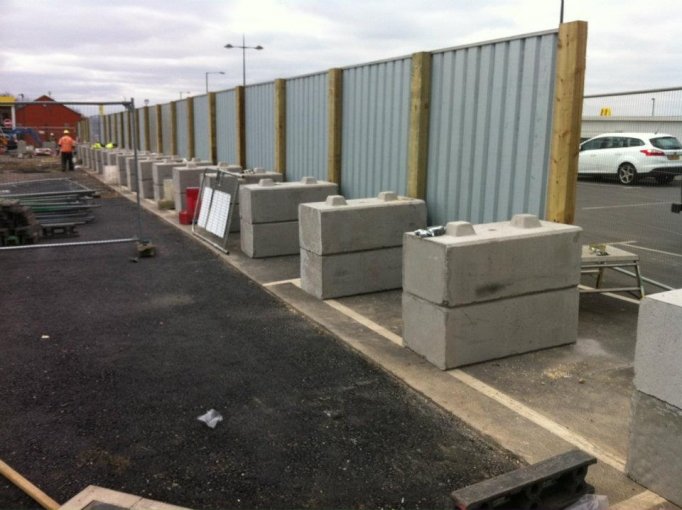 Kentledge blocks ballasting a run of site hoarding panels across a car park