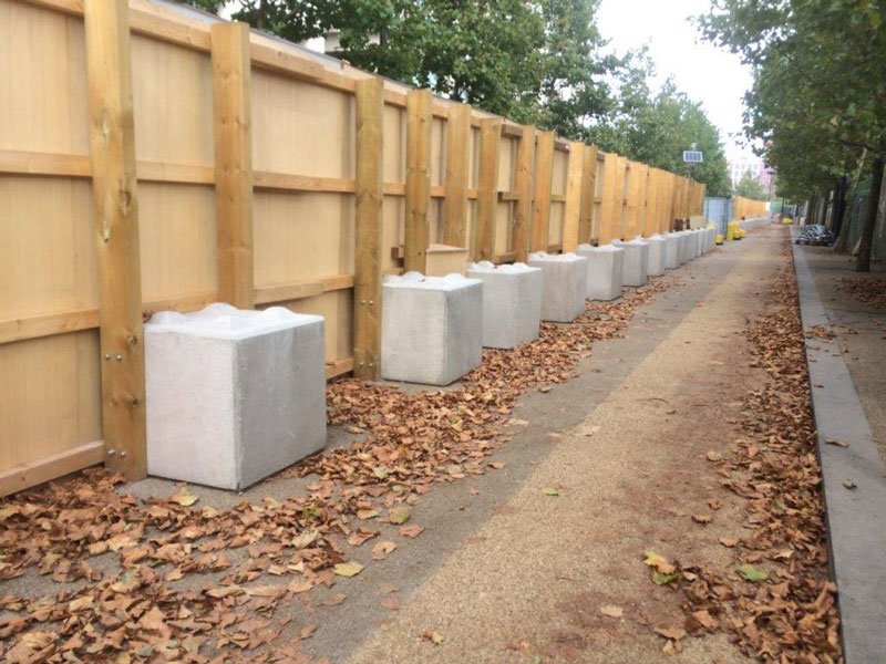 Kentledge blocks ballasting a timber hoarding along a leaf-strewn footpath