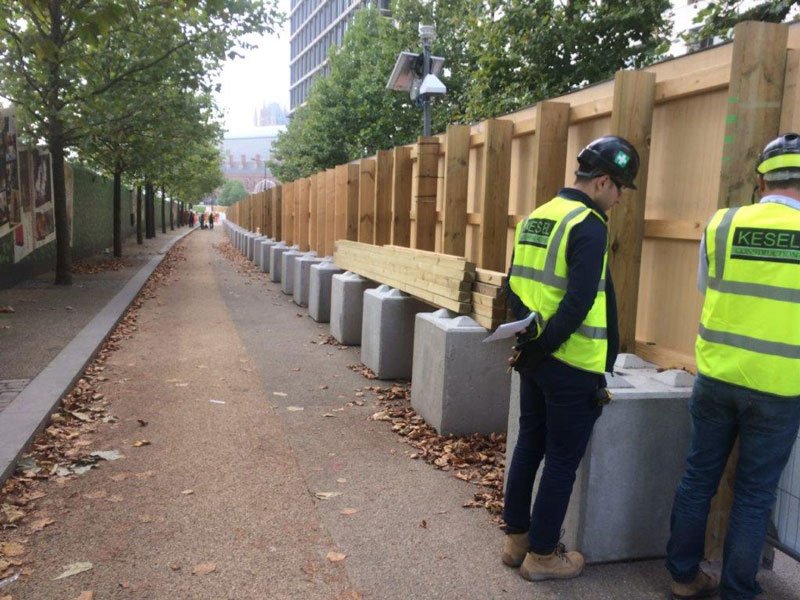 Workers installing precast blocks as ballast feet along a timber site hoarding
