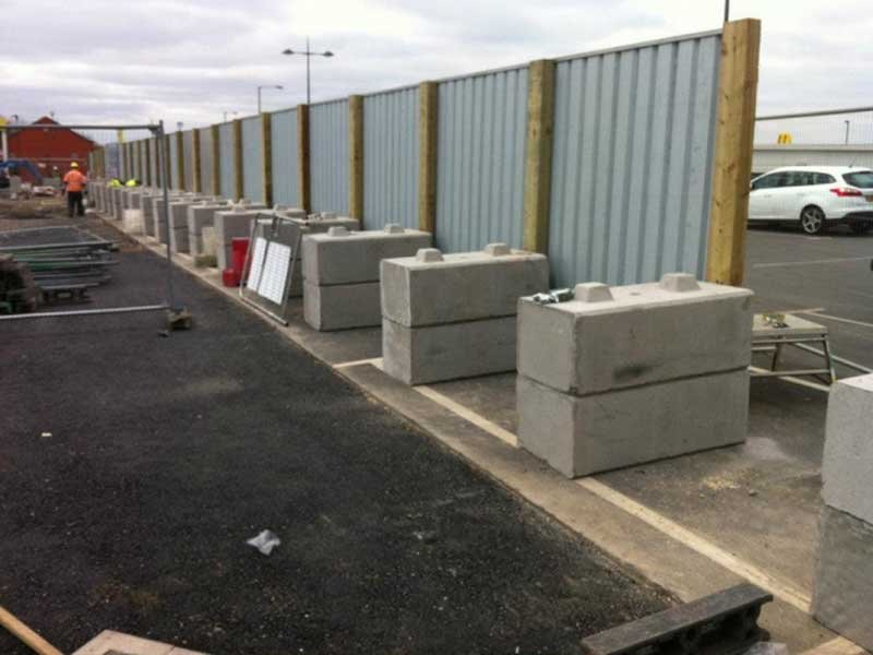 Concrete ballast blocks holding steel hoarding panels along a car park edge