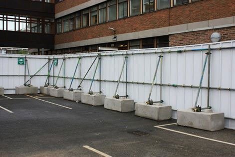 Site hoarding panels braced on concrete ballast blocks around a car park
