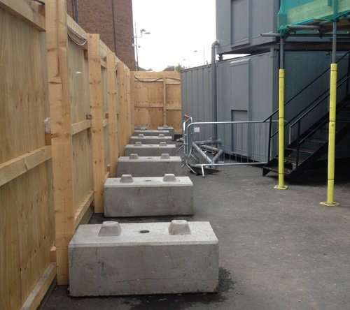 Row of concrete blocks ballasting timber hoarding along a narrow service alley