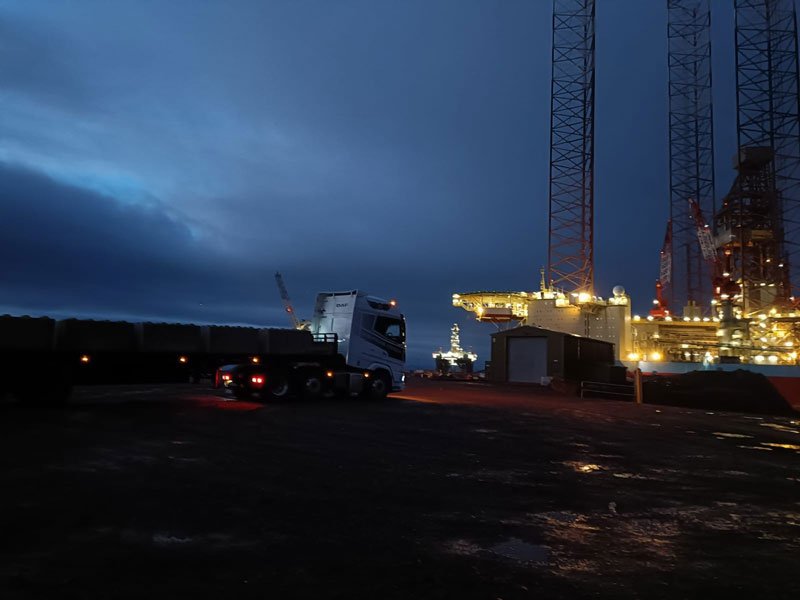 Lorry delivering blocks at a port at dusk with a lit rig beyond