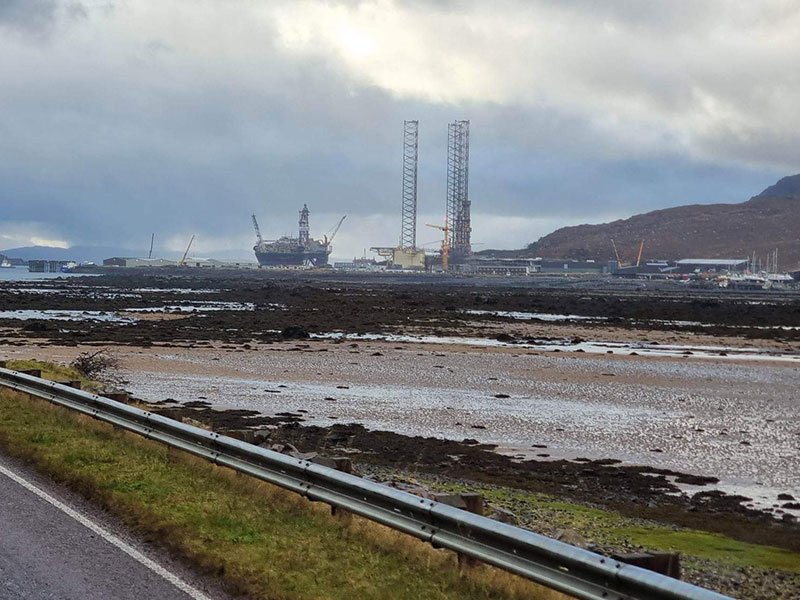 Jack-up oil rigs moored offshore seen across mudflats from a coastal road