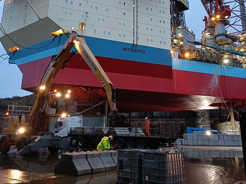 Excavator and lorry unloading concrete blocks beneath a floodlit rig hull