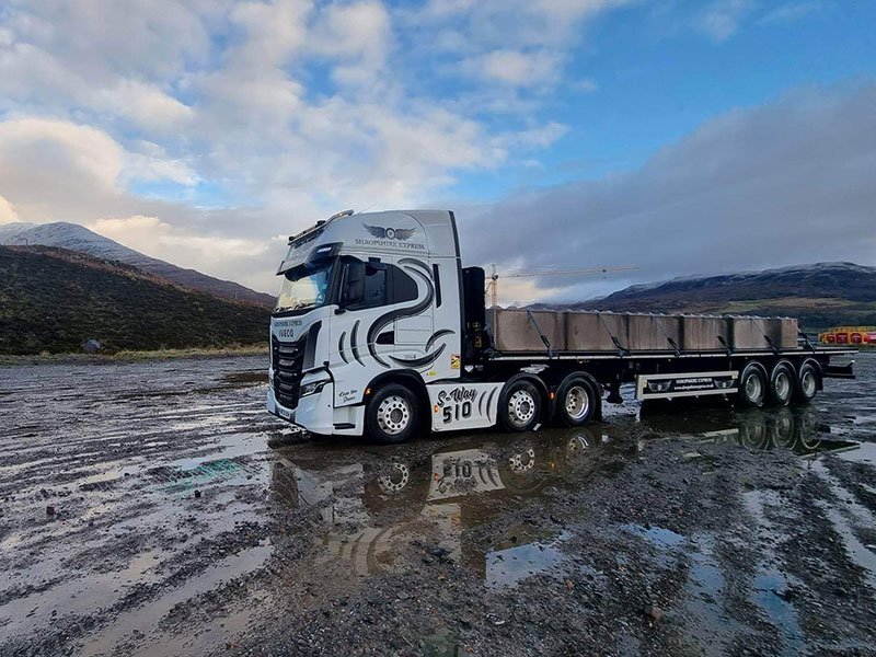 Articulated delivery lorry parked on a muddy coastal yard below the hills