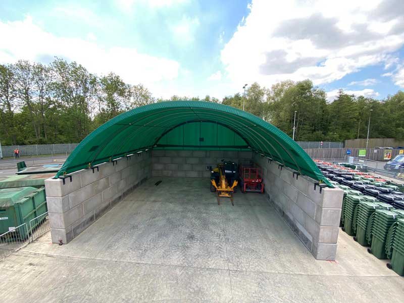 Green fabric-covered store on Legato block walls beside stacked wheelie bins