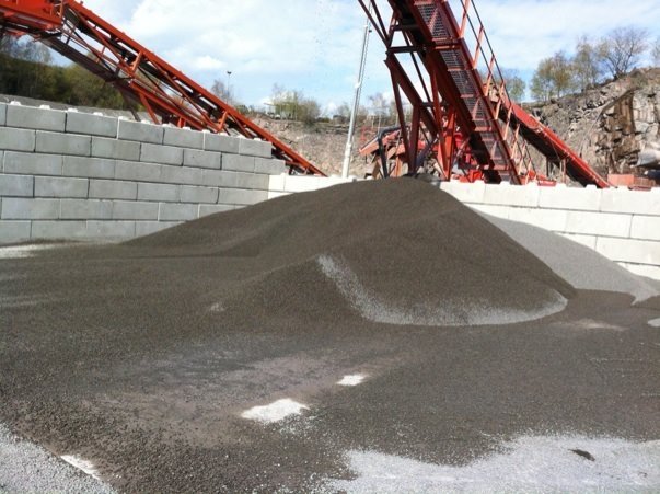 Fine aggregate stockpiled in a bay formed by interlocking blocks, conveyor behind
