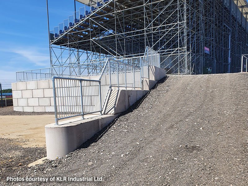 Gravel ramp climbing beside an interlocking block wall under a scaffolded grandstand