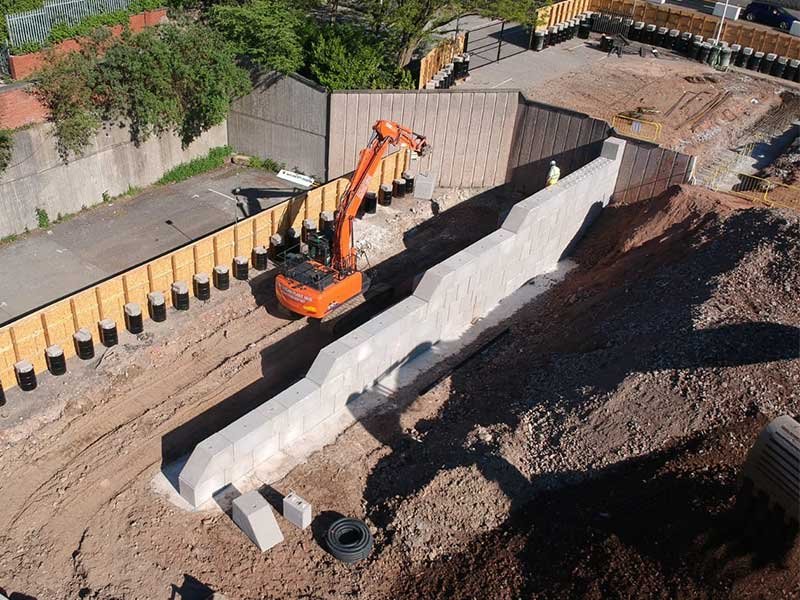 Excavator placing interlocking blocks in a trench beside a timber piled fence