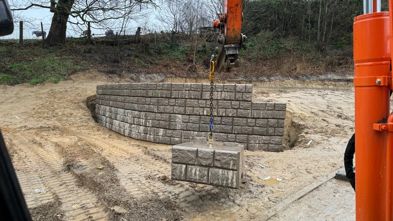 Excavator lowering a cobblestone-finish block onto a curved retaining wall