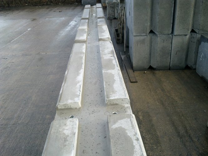 Long run of temporary vertical concrete barriers on a wet yard beside stacked units