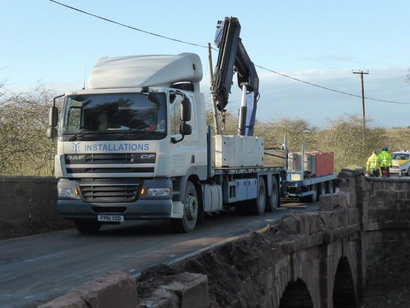 Lorry mounted crane delivering barriers across a narrow stone bridge under repair