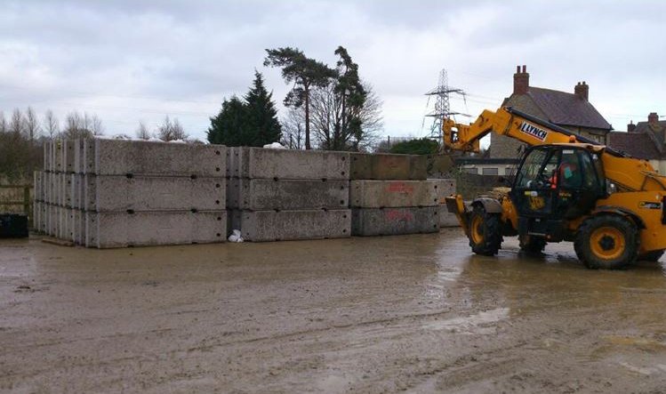Telehandler moving stacked concrete barrier units across a muddy flood defence site