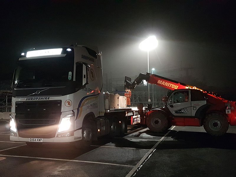 Telehandler offloading barriers from an articulated lorry under floodlights at night