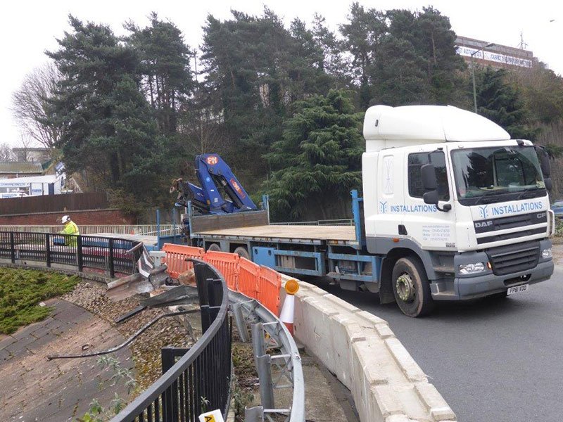 Crane lorry installing a concrete barrier along a roadside above railings
