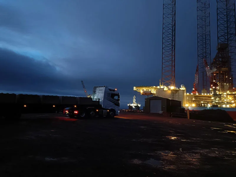 Lorry delivering concrete blocks at a port at dusk with lit rigs beyond