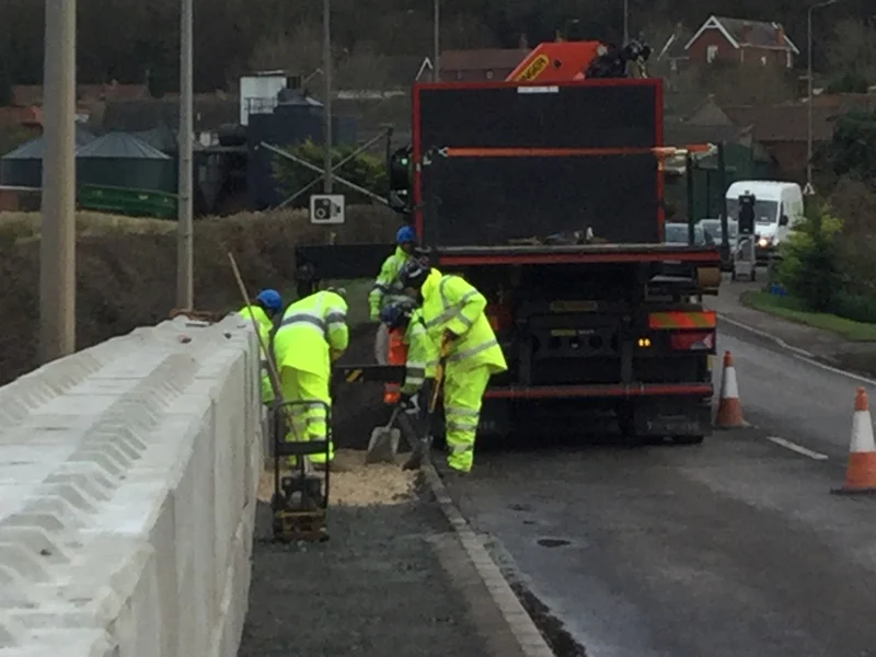 Workers in hi-vis bedding a precast concrete flood protection wall beside a road