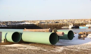 Large green pipes laid out on a shoreline beside harbour piling