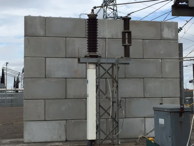 Interlocking concrete block blast wall shielding substation switchgear beneath overhead power lines
