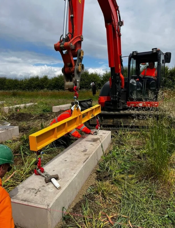 Excavator with a lifting beam placing a solar ballast block as a worker guides it