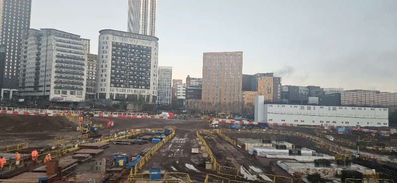 Cleared city-centre construction site with tower blocks and plant behind hoardings
