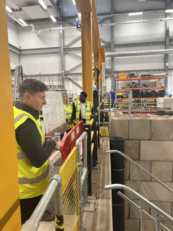 Man in a hi-vis vest briefing visitors beside a stone-faced block wall indoors